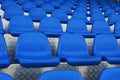 A close-up photograph of empty blue plastic stadium seats arranged in symmetrical rows on a metallic, textured floor