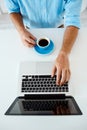 Close up picture of young businessman's hands sitting at table typing on laptop holding coffee cup. White modern office