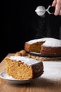 Close-up of piece of sponge cake with sugar on wooden table, in the background carrot sponge cake defocused, with hand pouring sug
