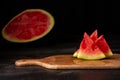 Close-up of pieces of watermelon on board, on dark wooden table, with half a watermelon in the background, black background, Royalty Free Stock Photo