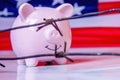 Close up pink piggy bank wrapped in barbed wire against United States national flag as symbol of economic warfare, sanctions and