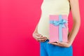 Close up of pink spotted gift box in pregnant woman`s hand wearing colorful home clothing at pink background. Waiting for a baby