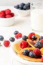 Close-up of plate of caramel pancakes with blueberries and raspberries with bowls and glass of milk, on white table, Royalty Free Stock Photo