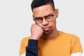 Close-up poratrait of tired Afro male leans on hand, has displeased expression, wearing sweaterand eyewear, posing in studio.