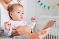 Close up portrait of a amazement baby sitting in mother`s arms and looking at the smartphone that she uses