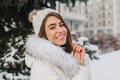 Close-up portrait of amazing girl in white knitted hat with snowflakes on hair smiling on street. Magnificent european