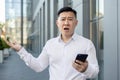 Close-up portrait of an angry young Asian man in a white shirt standing outside an office building, holding a phone and Royalty Free Stock Photo