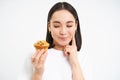 Close up portrait of asian woman with pensive smiling face, holding cupcake, thinking of bakery pastry, white background