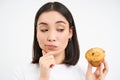 Close up portrait of asian woman with pensive smiling face, holds cupcake, thinks to eat pastry, white background