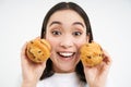 Close up portrait of asian woman, shows two cupcakes near face and smiles, likes pastry, enjoys eating bakery, white Royalty Free Stock Photo