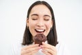 Close up portrait of asian woman smiles, eats chocolate cupcake with pleased face, isolated on white background Royalty Free Stock Photo