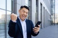 Close-up portrait of an Asian young business man standing in a suit near an office building, holding a phone in his Royalty Free Stock Photo