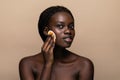 Close up portrait of an attractive african american woman removing makeup with sponge isolated on beige background