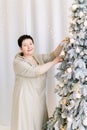 Close up portrait of attractive mature Caucasian woman, wearing elegant beige dress, decorating Christmas tree by Royalty Free Stock Photo