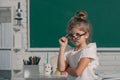 Close-up portrait of attractive small little cheerful girl sitting on table desktop in class room indoors. Little funny