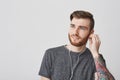 Close up portrait of attractive tattooed european man with beard and fashionable hairstyle plugging ear with earphone Royalty Free Stock Photo