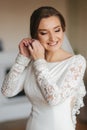 Close up portrait of beautiful bride standing by the window at home and tying on earrings. Charming bride in white Royalty Free Stock Photo