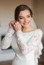 Close up portrait of beautiful bride standing by the window at home and tying on earrings. Charming bride in white Royalty Free Stock Photo