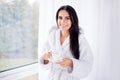 Close up portrait of beautiful brunette girl in bathrobe standing near window. She is smiling and holding a cup of coffee. Nice b