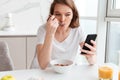 Close-up portrait of beautiful girl chatting on mobile phone while eating cornflakes at the kitchen table Royalty Free Stock Photo