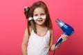 Close up portrait of beautiful little smiling girl with hair dryer and brush in hands, posing isolated over rosy studio wall with Royalty Free Stock Photo