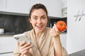 Close up portrait of beautiful young woman in the kitchen, holding tomato and smartphone, buys fresh vegetables online Royalty Free Stock Photo