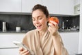 Close up portrait of beautiful young woman in the kitchen, holding tomato and smartphone, buys fresh vegetables online Royalty Free Stock Photo