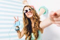 Close-up portrait of blissful girl in sunglasses and trendy bracelets posing with peace sign in studio. Charming young