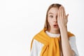 Close up portrait of blond girl covering half of face, looking with one eye and gasping, saying wow, staring surprised Royalty Free Stock Photo