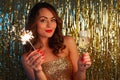 Young woman drinks champagne and holding a sparkler on a bright gold background, selective focus