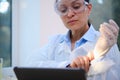 Close-up female scientist in white lab coat and safety goggles works on digital tablet and puts on protective gloves