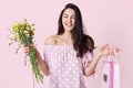 Close up portrait of cheerful female recieves present for birhday party, holds bag and flowers, looks happily, wears in dress,