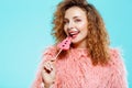 Close up portrait of cheerful smiling beautiful brunette curly girl in pink fur coat eating lollipop over blue Royalty Free Stock Photo