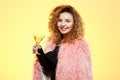 Close up portrait of cheerful smiling beautiful brunette curly girl in pink fur coat holding cocktail glass over yellow Royalty Free Stock Photo