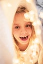 Portrait of a cheerful 6-year-old girl hiding in a white blanket and festive garlands