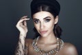 Glam dark-haired tattooed model with beautiful make up and smooth hair wearing black pillbox hat and luxurious gem necklace