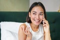 Close up portrait of cute asian girl in bed, talking on mobile phone with happy smiling face. Woman waking up and making Royalty Free Stock Photo