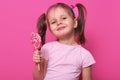 Close up portrait of cute child wearing rose casual t shirt, happy little girl holding big sugar lollipop, has gladness facial Royalty Free Stock Photo