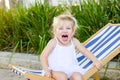 Close up portrait of cute emotional blondy toddler girl in white dress sitting on the deckchair and yelling. City park recreation Royalty Free Stock Photo