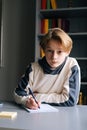 Close-up portrait of cute pupil boy doing homework writing to notebook with pen sitting at desk Royalty Free Stock Photo