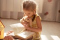 Close up portrait of cute toddler girl with dark hair reading book for her soft toy while sitting on floor in her bedroom, reads Royalty Free Stock Photo