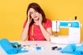 Close up portrait of dark haired young woman works as seamstress, sews new dress for her daughter, looks tired, sits near sewing