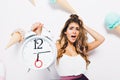 Close-up portrait of dismayed brunette woman in stylish outfit touching hair and holding big clock. Stunning young woman