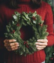 Woman in knitted sweater holds green Christmas wreath in hands.