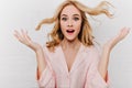 Close-up portrait of fair-haired girl in pink attire expressing amazement on white background. Surprised blonde lady in