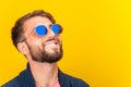 Close up portrait of funky young bearded hipster man in blue sunglasses smiling brightfully on a yellow background. Copy