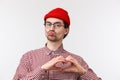 Close-up portrait funny caucasian male with geeky look, wear glasses and red beanie, pouting for kiss, showing heart