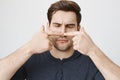 Close-up portrait of funny guy with cute haircut touching his nose with both index fingers and looking at it, standing Royalty Free Stock Photo