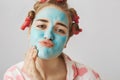 Close-up portrait of funny white woman with hair-curlers, wearing nightwear and folding lips while applying face mask Royalty Free Stock Photo