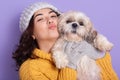 Close up portrait of girl wearing yellow knitted stylish outfit showing kiss gesture, posing with her puppy. Indoor portrait of